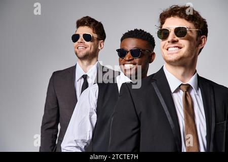 amici allegri e variegati in un abbigliamento casual con occhiali da sole in posa su sfondo grigio Foto Stock