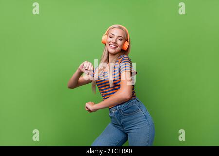 Foto di una donna stravagante che indossa una t-shirt a righe che balla musica pop con cuffie arancioni e che vibra la nostalgia isolata su sfondo verde Foto Stock