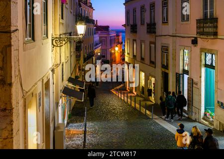 Serata nel quartiere collinare di Alfama mentre i turisti e i portoghesi camminano per le strette strade acciottolate passando davanti a negozi illuminati e caffetterie, a Lisbona in Portogallo. Foto Stock