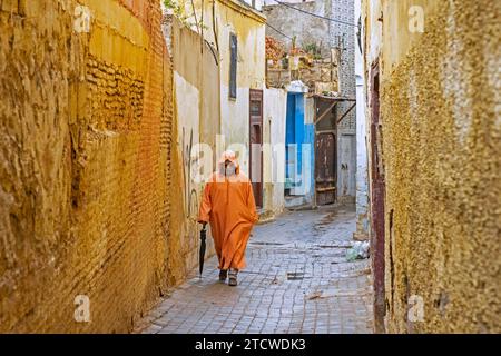 Uomo musulmano che indossa djellaba/jillaba arancione in un giorno di pioggia nel vicolo della medina gialla della città di Meknes, Fez-Meknes, Marocco Foto Stock