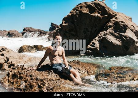 Uomo seduto sulle rocce nell'acqua alla spiaggia del Monte Maunganui, nuova Zelanda. Foto Stock