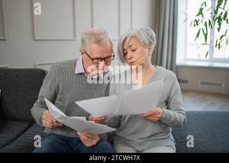 Triste stanco, deluso, coppia di anziani di mezza età siedono con un documento cartaceo. Infelice anziano maturo donna che legge banconote che gestiscono le finanze bancarie calcolano le tasse pianificazione prestito pagamento pensione Foto Stock