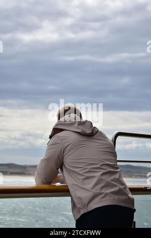 Giovane uomo con pantaloni neri e giacca grigia con cappello appoggiato su un corrimano su una nave che naviga in mare vicino alla costa Foto Stock