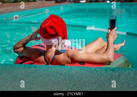 Una donna felice in bikini blu, un cappello di Babbo Natale rosso e bianco e occhiali da sole si posa in piscina in un cerchio gonfiabile con un motivo anguria, reggendo Foto Stock