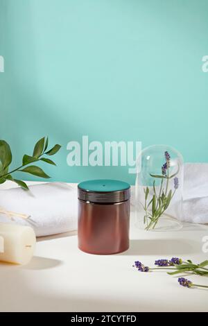 Un barattolo marrone di scrub è esposto sul tavolo con asciugamani di cotone, foglie verdi, una candela e fiori di lavanda. Cura della pelle con prodotti contenenti na Foto Stock