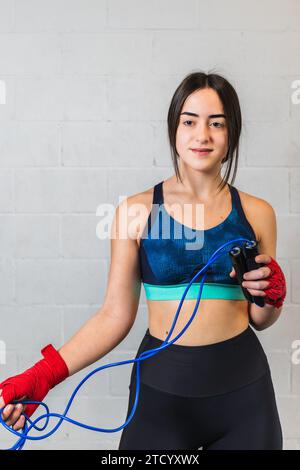 Foto verticale bella ragazza adolescente caucasica, vestita con abiti sportivi, con bende rosse sulle mani e una corda blu. In una palestra di boxe su mattoni bianchi c Foto Stock