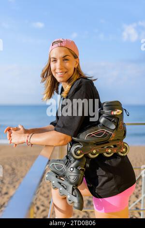 Ritratto di una giovane donna caucasica vestita di casual con pattini a rotelle in linea appoggiati su una capanna bagnino che ringhiera in spiaggia sotto il cielo blu Foto Stock
