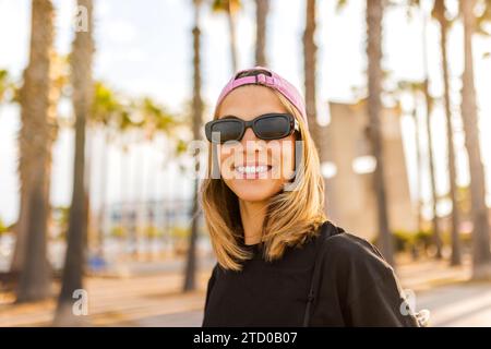 Ritratto di una giovane donna sorridente che indossa occhiali da sole neri e maglietta con berretto rosa su sfondo sfocato nel parco durante il fine settimana Foto Stock