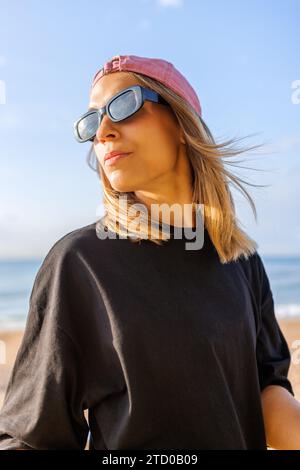Giovane ed elegante donna bionda che indossa occhiali da sole e berretto che guarda dietro le spalle mentre si trova in spiaggia contro il cielo blu durante il giorno di sole Foto Stock