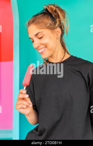 Ritratto di una bellissima giovane donna caucasica con ghiacciolo rosa mentre si trova contro la parete blu Foto Stock