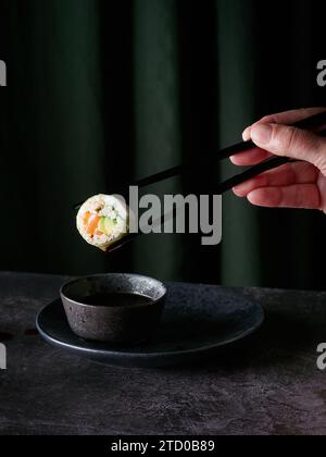 Sushi roll tenuto da una persona anonima utilizzando delle bacchette con una goccia di salsa pronta a cadere nella ciotola posta sul tavolo contro le tende verdi Foto Stock