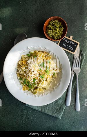 Scatto in alto di una deliziosa ciotola di pesto farfalle con formaggio grattugiato, su sfondo scuro con una piccola ciotola di erbe tritate e un'etichetta indi Foto Stock