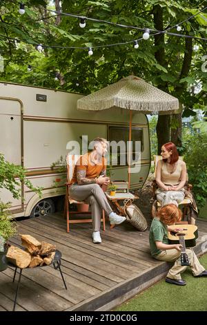 redhead boy che suona la chitarra acustica mentre i genitori sono seduti e parlano accanto al trailer di casa Foto Stock