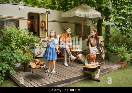 bambini che giocano a badminton e alla chitarra acustica vicino ai genitori seduti accanto a una casa di roulotte moderna Foto Stock