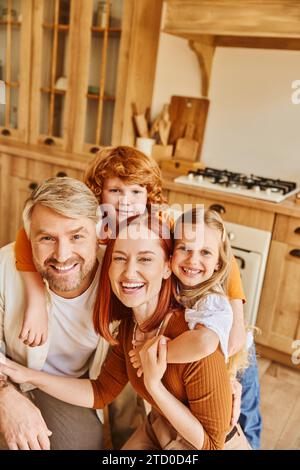 genitori e bambini felicissimi che abbracciano e guardano la macchina fotografica in una cucina accogliente, momenti preziosi Foto Stock