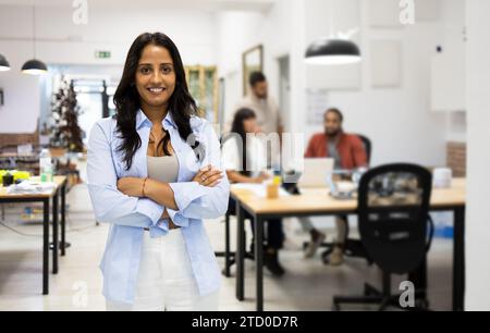 Ritratto di una donna d'affari latina sorridente e sicura di sé vestita di casual in piedi con le braccia incrociate su sfondo sfocato in un ufficio creativo Foto Stock