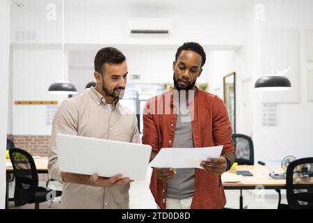 Colleghi maschili multirazziali che discutono di rapporti su documenti e laptop mentre si trovano in un ufficio creativo Foto Stock