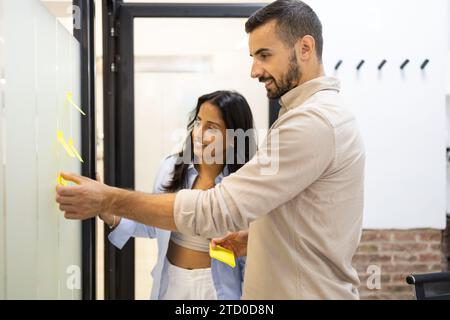 Colleghi multirazziali che indossano abiti casual appiccano appunti adesivi su una parete di vetro e discutono le strategie aziendali durante la riunione in un ufficio creativo Foto Stock