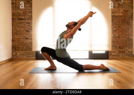 Un uomo esegue uno yoga su un tappetino in una stanza con pavimenti in legno duro e pareti in mattoni a vista, illuminata da luce naturale. Foto Stock