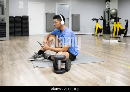Un giovane concentrato siede su un tappetino in palestra, controlla il suo smartphone mentre indossa le cuffie, con l'attrezzatura per l'esercizio nelle vicinanze. Foto Stock