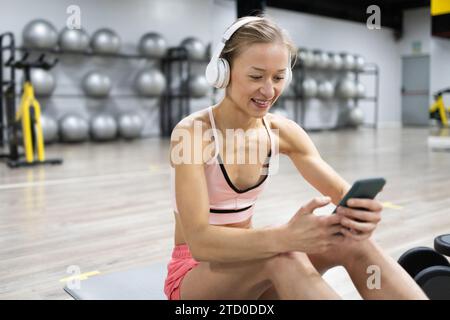 Una giovane donna incentrata sul fitness si prende una pausa, interagendo felicemente con il suo smartphone in palestra, indossando cuffie e abbigliamento da allenamento. Foto Stock