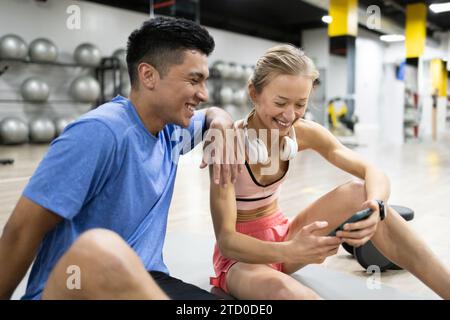 Due appassionati di fitness fanno una pausa in palestra, ridendo insieme mentre guardano uno smartphone con le palle da ginnastica sullo sfondo. Foto Stock