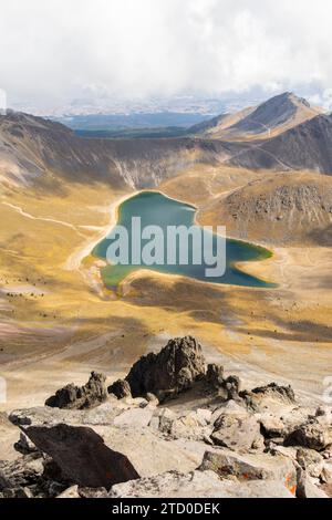 Una vista panoramica di un sereno lago cratere incastonato tra i paesaggi scoscesi del Nevado de Toluca, con sfumature vivaci che si riflettono sull'acqua. Foto Stock