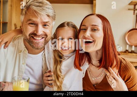 genitori eccitati con una figlia carina che guarda la macchina fotografica in cucina a casa, interazione con la famiglia Foto Stock