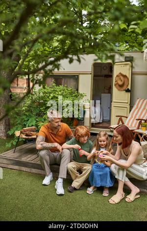 donna che mostra come giocare con le bolle di sapone ai bambini seduti accanto a una casa di roulotte moderna Foto Stock