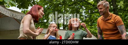 genitori felici che guardano i bambini carini che soffiano bolle di sapone vicino a roulotte, all'aperto, banner Foto Stock