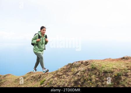Vista a tutto il corpo di una giovane escursionista in un impermeabile verde con zaino che sale sulla vetta della montagna contro il cielo durante le vacanze del fine settimana Foto Stock