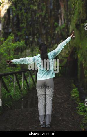 Vista sul retro di una giovane donna felice con le braccia aperte lungo un sentiero escursionistico vicino alla scogliera verde durante il fine settimana Foto Stock