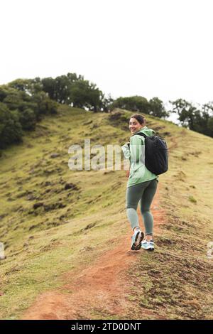 Vista laterale di una giovane e felice escursionista che indossa un impermeabile con zaino che cammina sulla montagna e guarda a spalla contro il cielo limpido Foto Stock