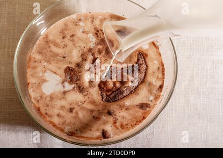 Vista dall'alto di Un flusso di latte versato in una ciotola di vetro ripiena di cioccolato posta sul tavolo Foto Stock