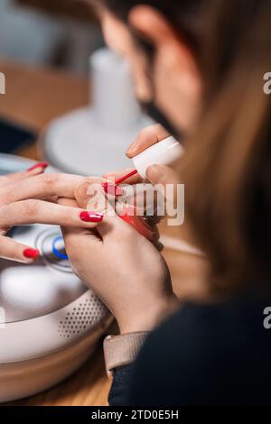 Dall'alto del prodotto estetista anonimo in maschera protettiva applicando smalto per unghie di clienti irriconoscibili presso un moderno salone di bellezza Foto Stock