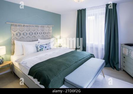Camera da letto accogliente ed elegante con letto bianco con accenti verdi scuro, eleganti tendaggi e pareti decorative. Foto Stock