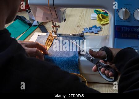 Mani precise di un sarto che cuciscono un pezzo di tessuto denim blu utilizzando una macchina da cucire bianca, con forbici e metro a nastro in uso. Foto Stock