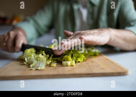 Tagliate le mani di un anonimo uomo anziano che taglia la lattuga a pezzi con il coltello su tagliere di legno mentre preparate l'insalata a casa Foto Stock