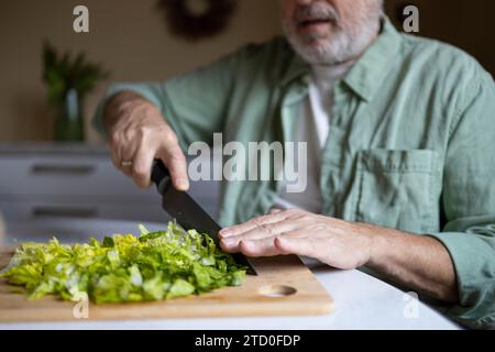 Tagliate le mani di un anonimo uomo anziano che taglia la lattuga a pezzi con il coltello su tagliere di legno mentre preparate l'insalata a casa Foto Stock