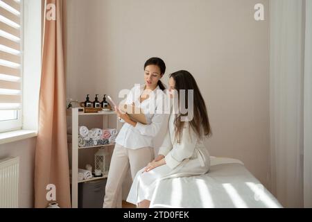 Massaggiatrice sicura di sé con appunti in uniforme e consulenza a una cliente seduta sul letto per la procedura terapeutica in salone Foto Stock