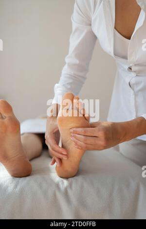 Ritaglia le mani di una terapista anonima che massaggia il piede del cliente sdraiato sul letto per alleviare il dolore premendo delicatamente i punti di pressione presso il salone termale Foto Stock