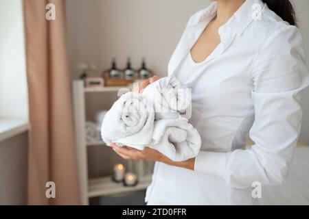 Ritaglia la mano di una massaggiatrice anonima in uniforme bianca in piedi e tieni asciugamani arrotolati puliti mentre lavori nel salone termale Foto Stock