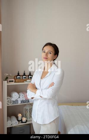 Ritratto di massaggiatrice sicura e positiva in uniforme bianca con le braccia incrociate sorridendo e guardando la macchina fotografica mentre si è a letto nella spa Foto Stock