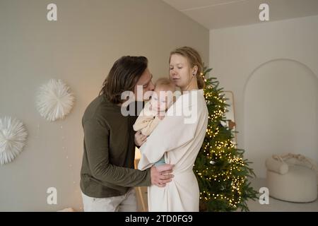 Una famiglia amorevole condivide un momento di tenerezza con il bambino vicino a un albero di Natale adornato di luci. Foto Stock