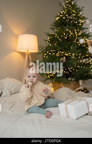 Un bambino carino, vestito in modo caldo, siede di fronte a un albero di Natale, con luci e regali intorno, godendosi l'atmosfera delle vacanze. Foto Stock