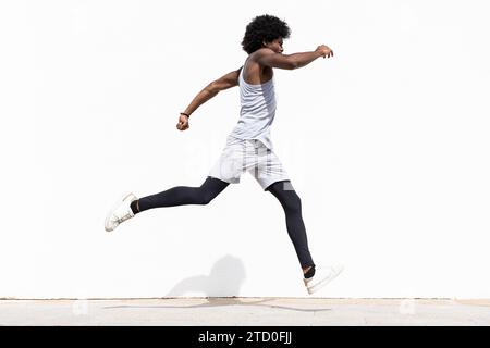 Vista laterale corpo completo di giovane atleta afro-americano attivo in abbigliamento sportivo bianco che corre contro parete bianca durante la giornata di sole Foto Stock
