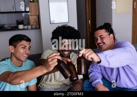 Multirazziale allegro e allegro giovane amico maschio in abiti informali brindando bottiglie di birra mentre si siede sul divano in soggiorno durante le vacanze del fine settimana Foto Stock
