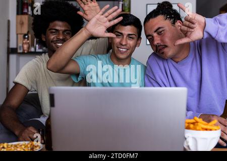 Allegri giovani amici maschi vestiti con le mani che saltano e gestiscono mentre videochiamano tramite computer wireless in salotto durante il tempo libero tim Foto Stock