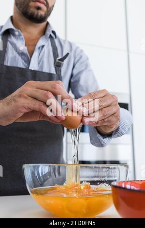 Ritaglia lo chef maschile barbuto anonimo in grembiule in piedi versando l'uovo rotto nella ciotola di vetro sul tavolo della cucina e preparando la colazione alla luce del giorno Foto Stock
