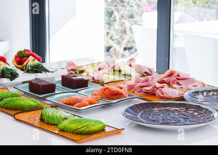 Alto angolo di appetitoso affettato di avocado salmone e prosciutto servito su piatti vicino al dessert e frutti di bosco messi sul bancone in una cucina leggera Foto Stock